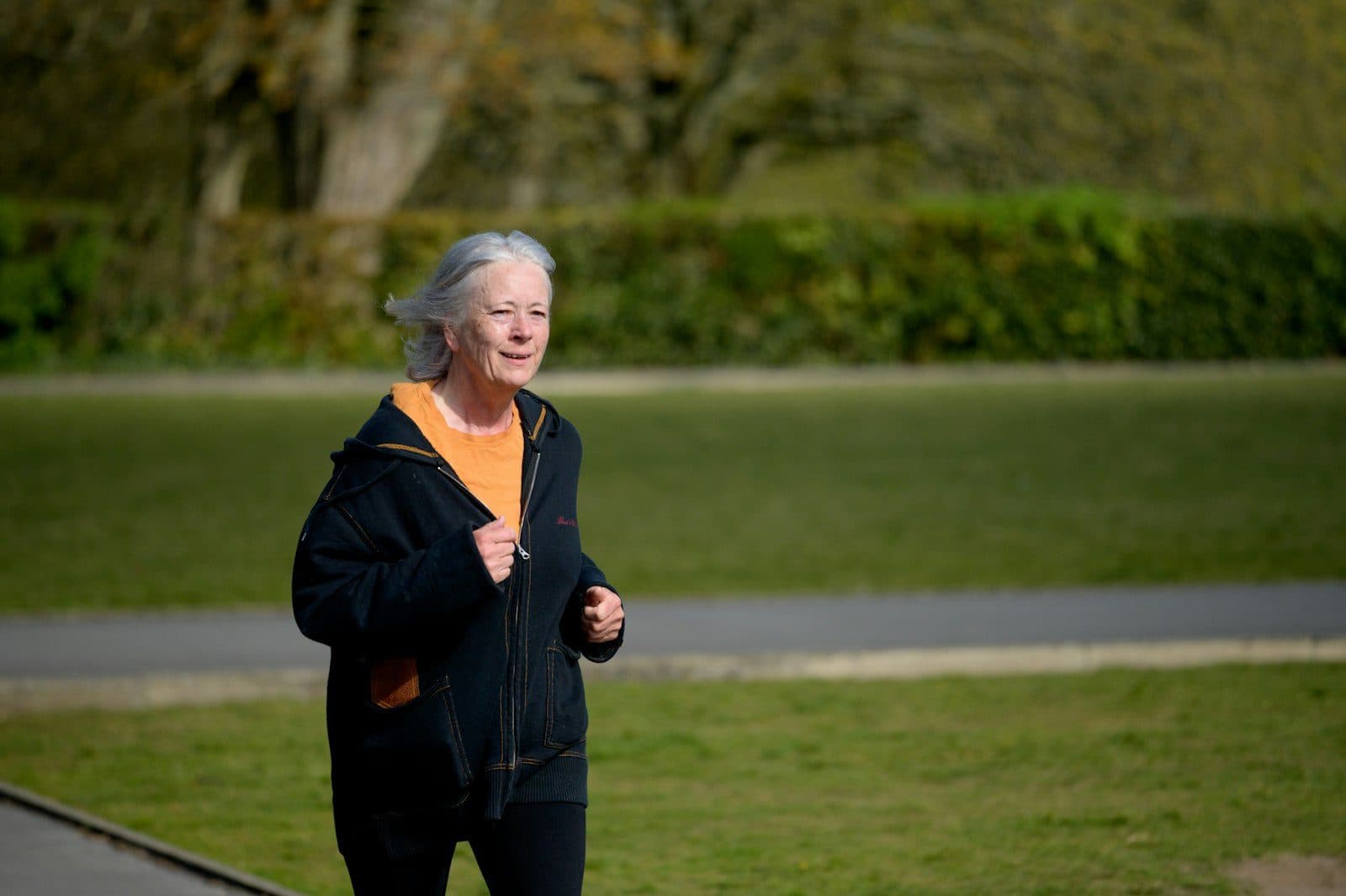 An older woman in a dark jacket jogging across a park