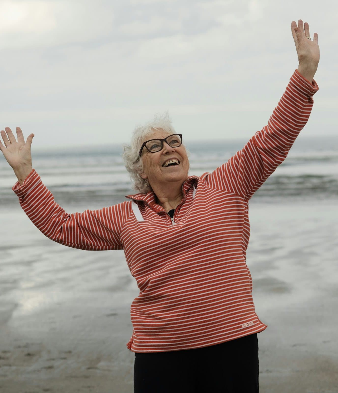 A smiling older woman in a coral striped top raising both arms on a beach